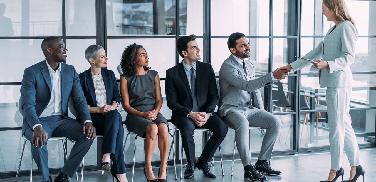 Line of job applicants wait in chairs while one shakes hands with someone.