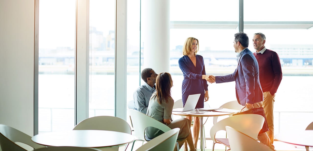 A group of 5 professionals sit in a glass-walled conference room, with two shaking hands.